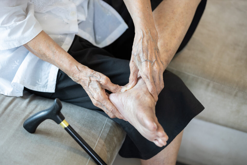 A close-up shot of an elderly person’s hands massaging their bare foot while sitting down. Their skin appears thin with prominent veins. A black walking cane rests on the sofa cushion next to them, suggesting mobility challenges.