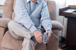 An elderly man holds his foot suffering from foot pain while sitting on a sofa in a room