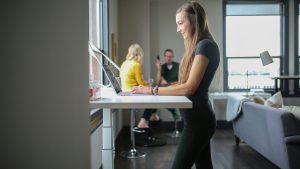 a young woman using a standing desk with coworkers visible in the background