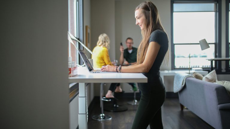 a young woman using a standing desk with coworkers visible in the background